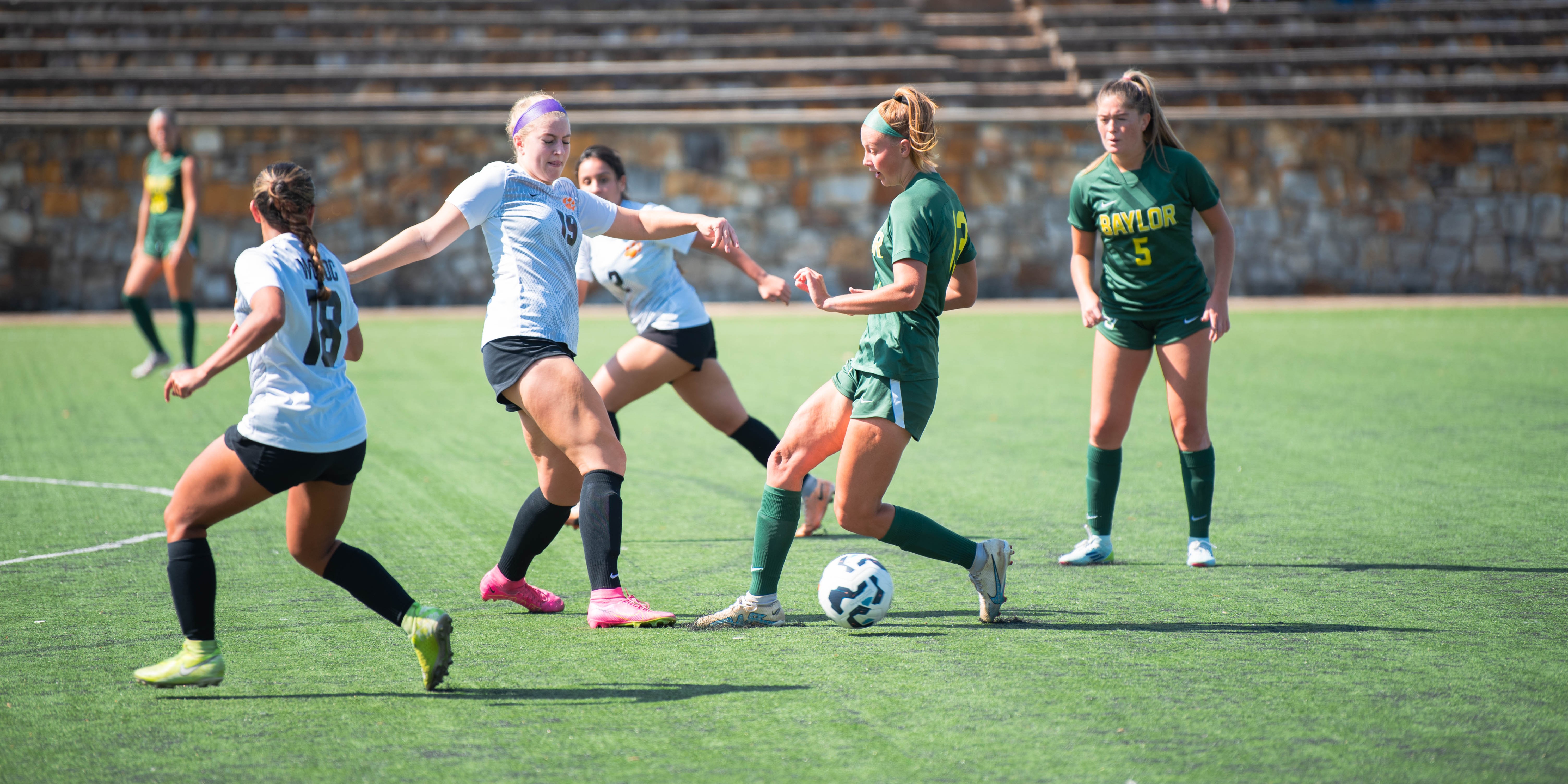 three womens soccer players going after ball on field on sunny day